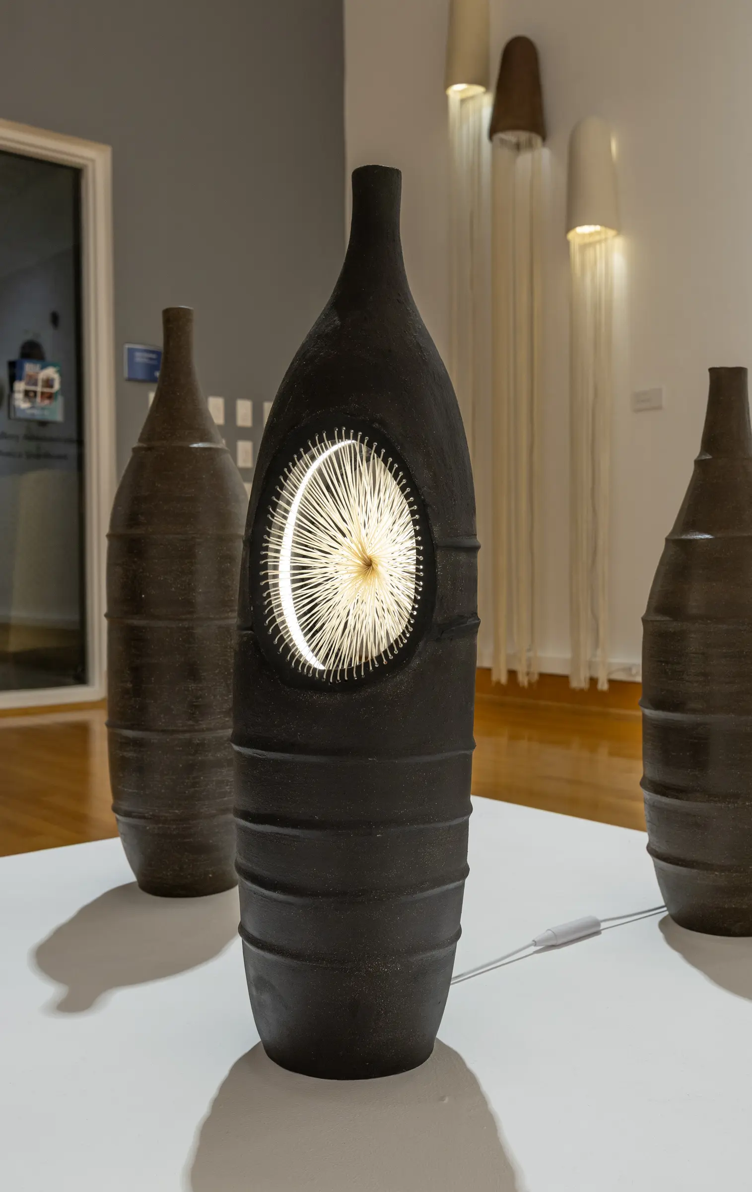 Tall black ceramic vessel illuminated through a woven circular opening